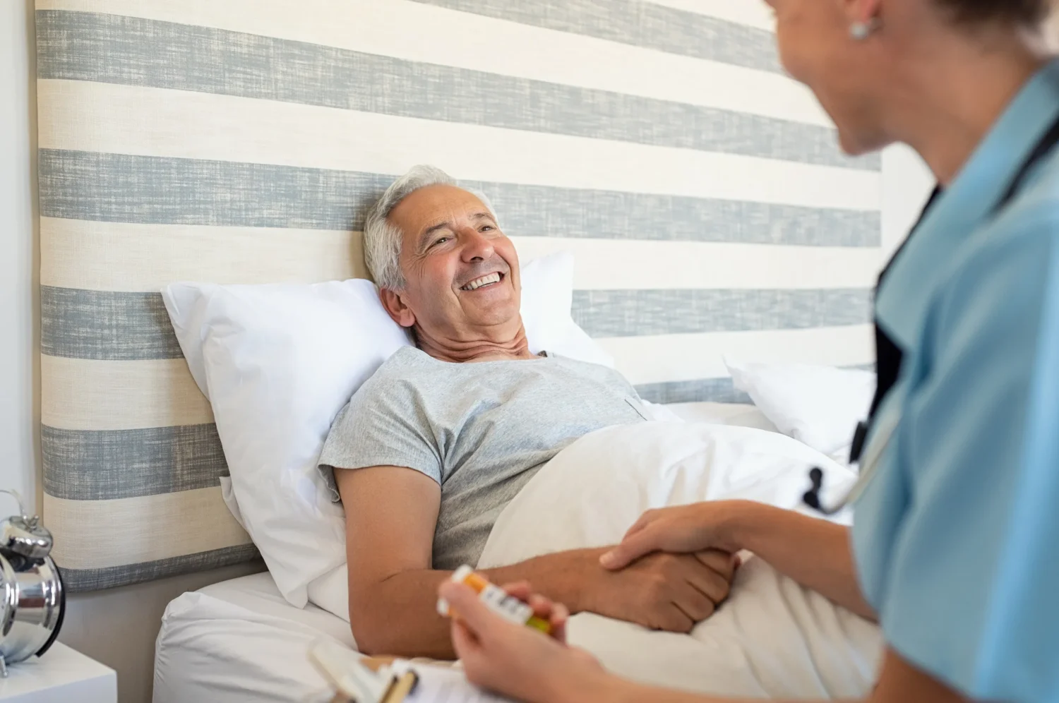 Nurse holds the hand of a patient lying in bed, discussing medication, in a bright, simple hospital room with a striped wall