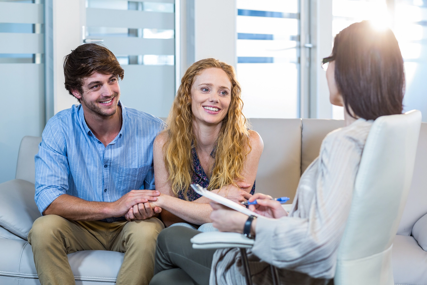 A couple sits on a sofa in a counseling session, holding hands while a therapist takes notes and engages with them