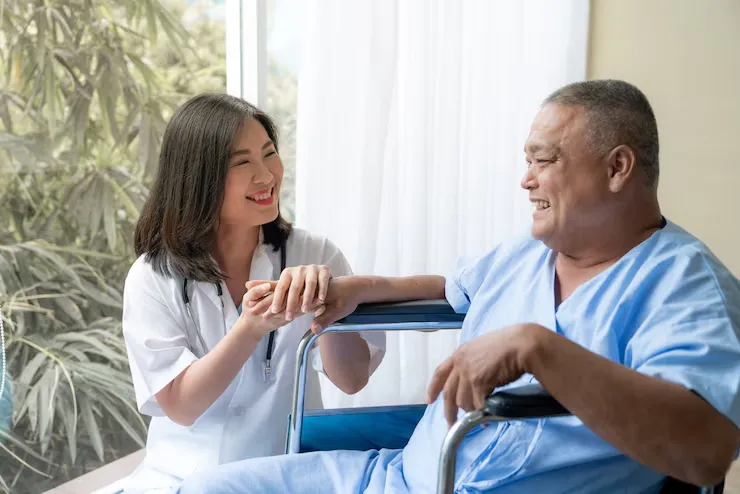 An Asian man in a wheelchair sits beside a nurse, who is engaged in conversation and offering care